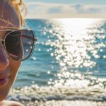 A woman wearing stylish polarized sunglasses on a sunny beach, with ocean waves reflecting bright light in the background