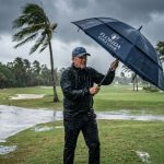 Golfer holding a large windproof golf umbrella on a Florida course during a heavy rainstorm with palm trees bending in the wind