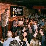 A packed comedy club in New York City during the NYC Comedy Festival with a comedian performing on a lit stage in front of a laughing audience.