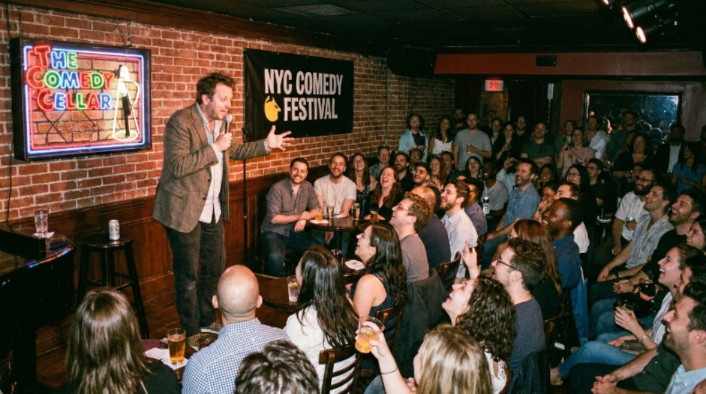 A packed comedy club in New York City during the NYC Comedy Festival with a comedian performing on a lit stage in front of a laughing audience.