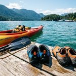 Neoprene kayaking shoes rest on a sunny dock next to a kayak.